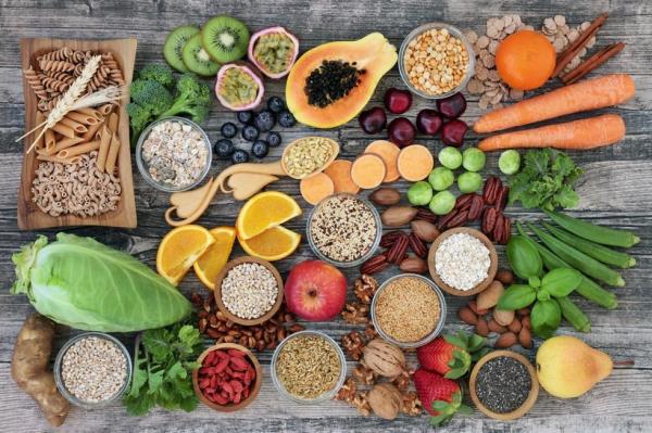 Whole foods on a wood table with fruit, vegetables, whole wheat pasta, legumes, cereals, nuts and seeds